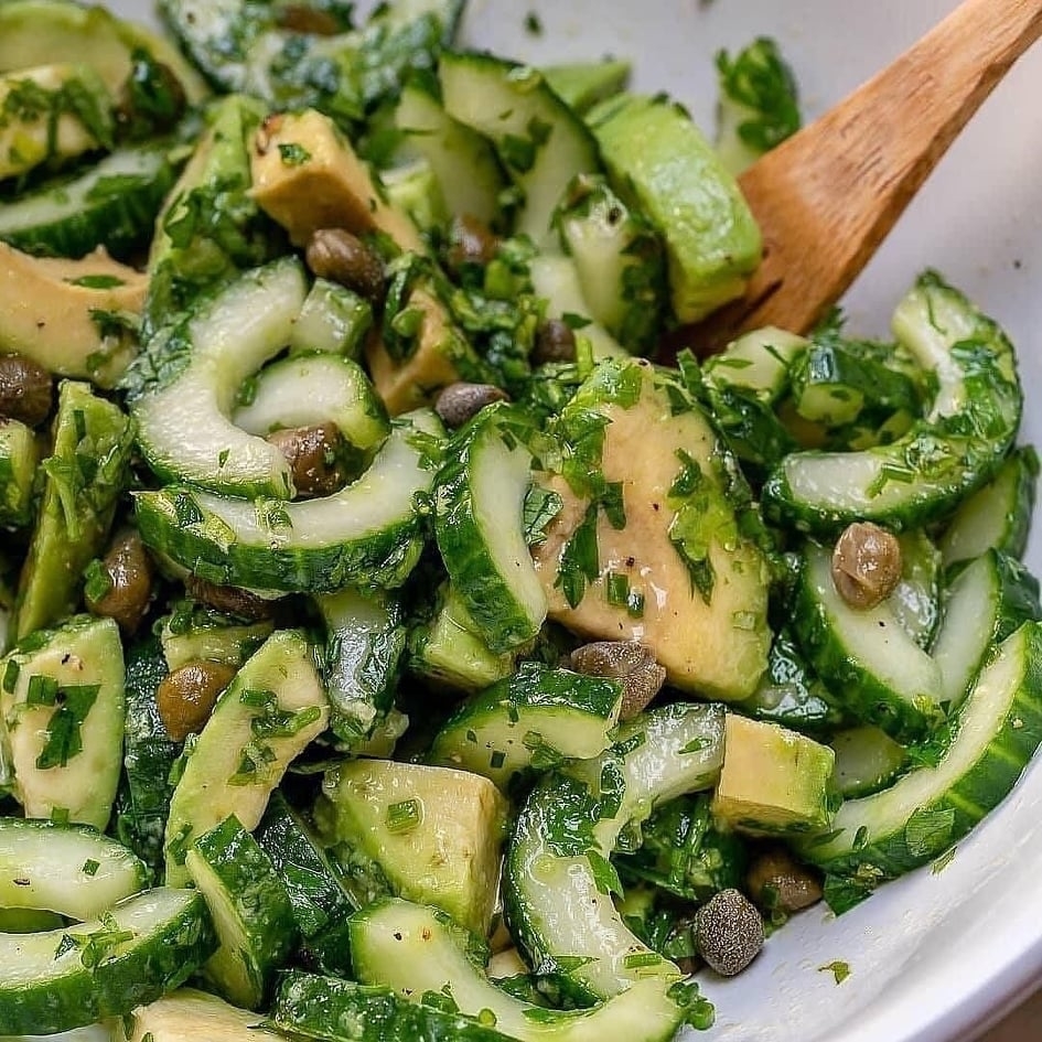A close-up of a fresh salad with sliced cucumbers, avocado, capers, and chopped herbs, mixed in a bowl with a wooden spoon