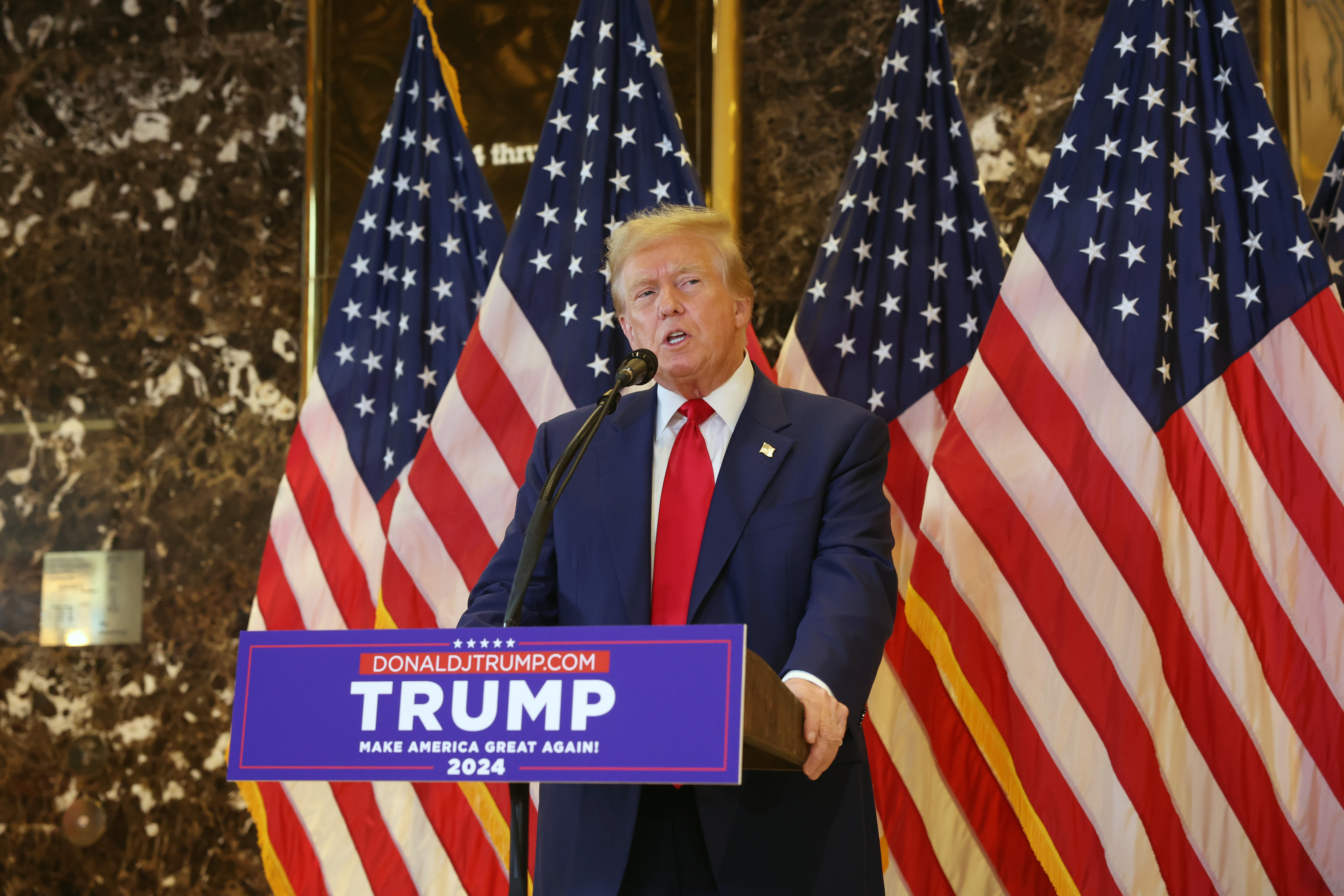 Donald Trump stands at a podium with "Trump 2024" signage, flanked by multiple American flags