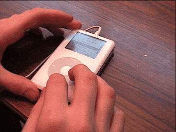 Hands holding and using a white iPod with a click wheel on a wooden table. The screen displays a menu with text options