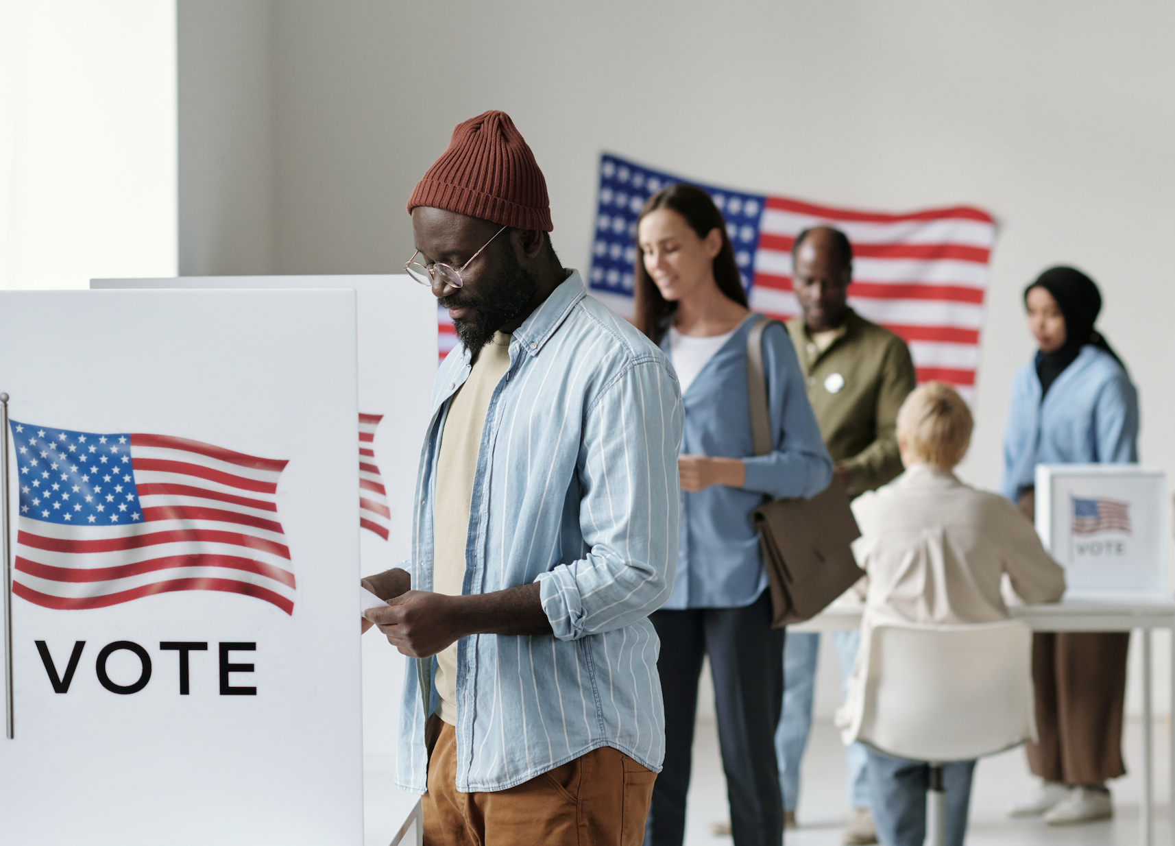 People voting in an election polling place with American flags in the background. A man in a beanie is looking at a paper while standing at a voting booth
