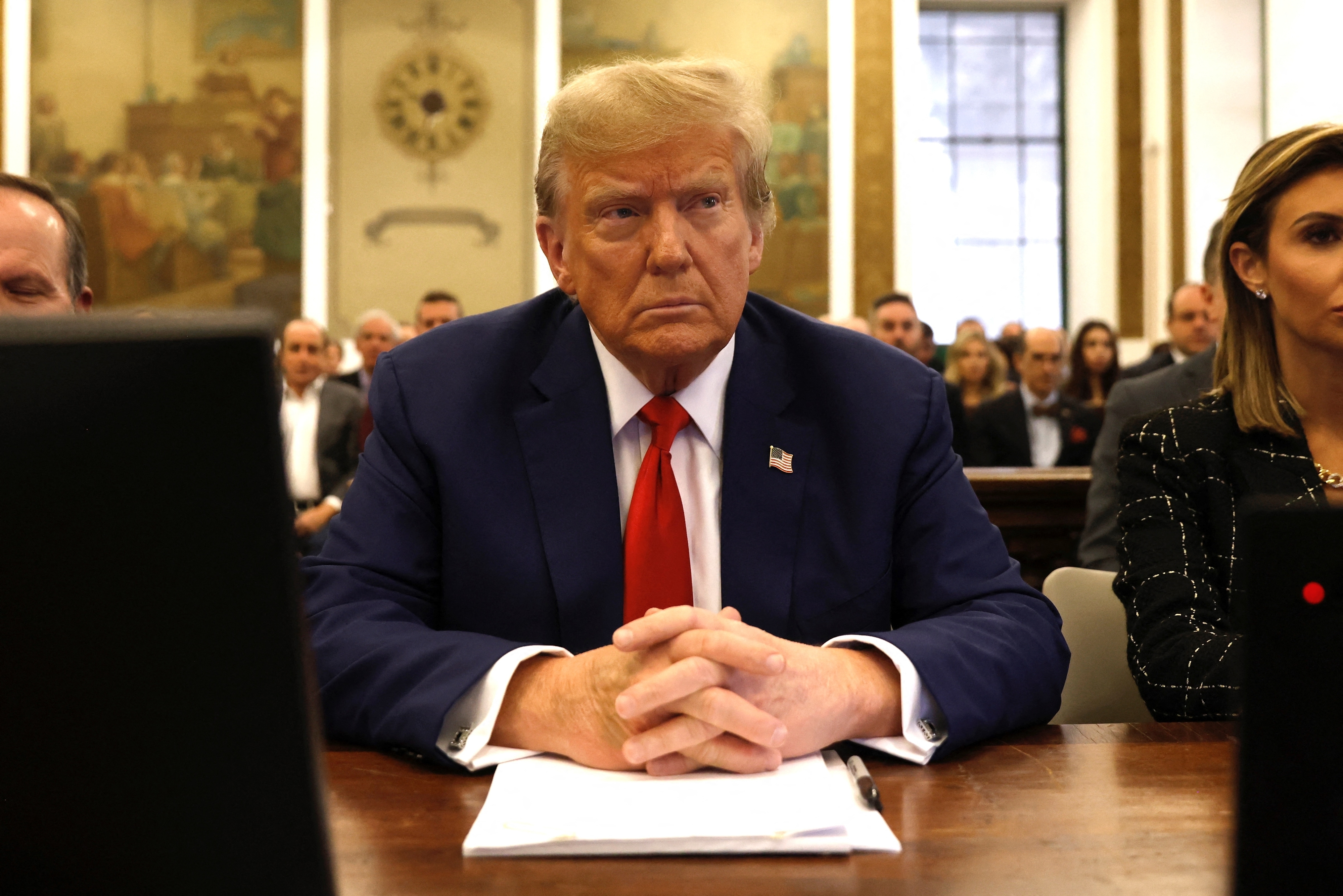 Donald Trump seated at a table, hands clasped, wearing a suit and tie, with people in the background inside a court
