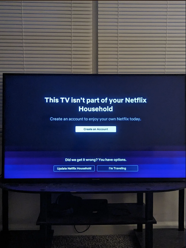 A TV screen displays text stating that the TV isn't part of the user's Netflix household, with options to create an account, update the household, or indicate traveling