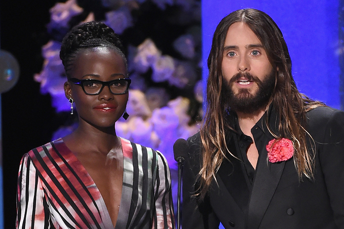Lupita Nyong'o in a stylish striped dress and Jared Leto in a black suit with a pink rose lapel pin, both speaking on stage