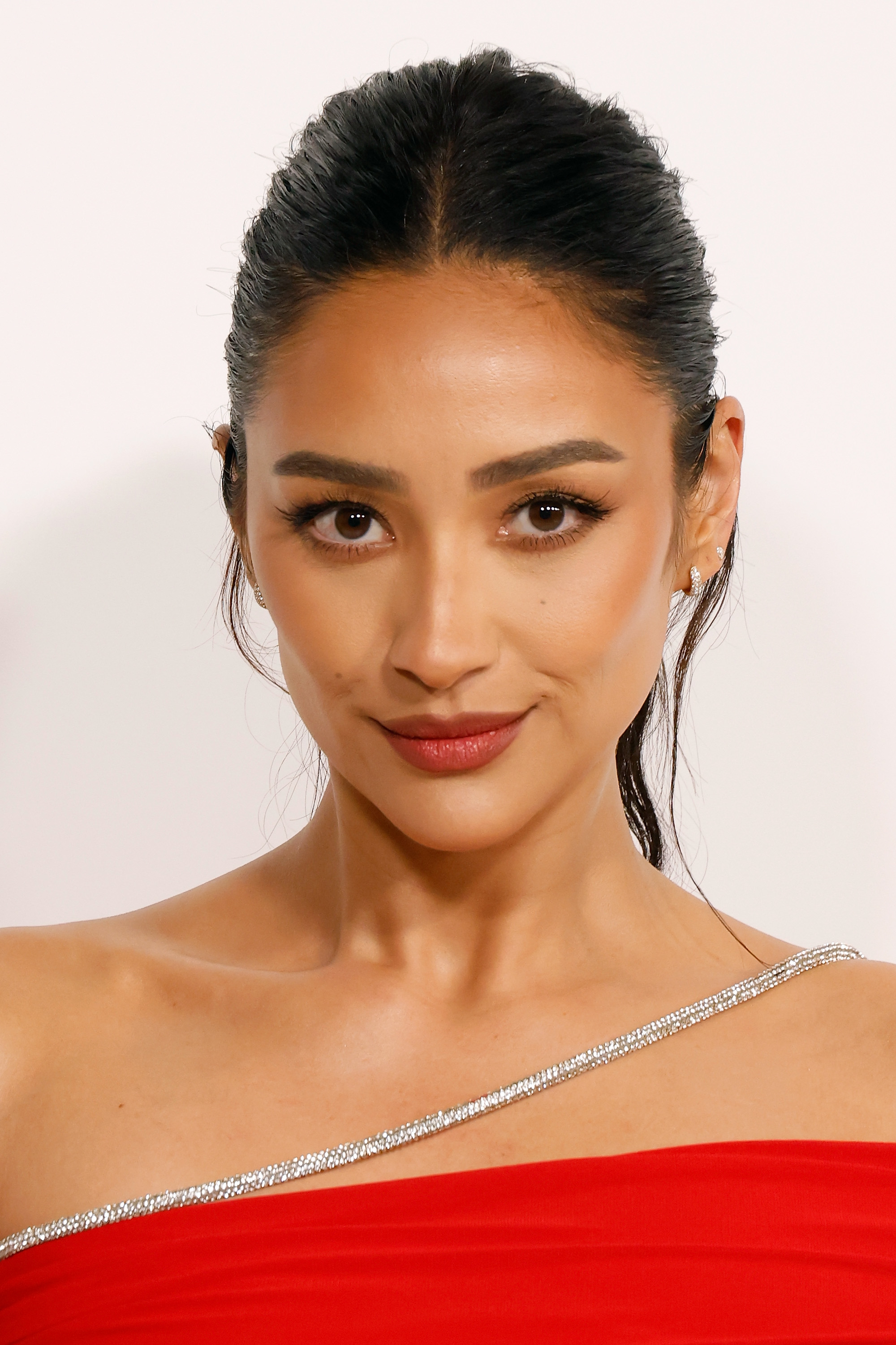 Shay Mitchell posing; she has a sleek hairstyle and wears an elegant, off-the-shoulder red dress with a sparkling trim
