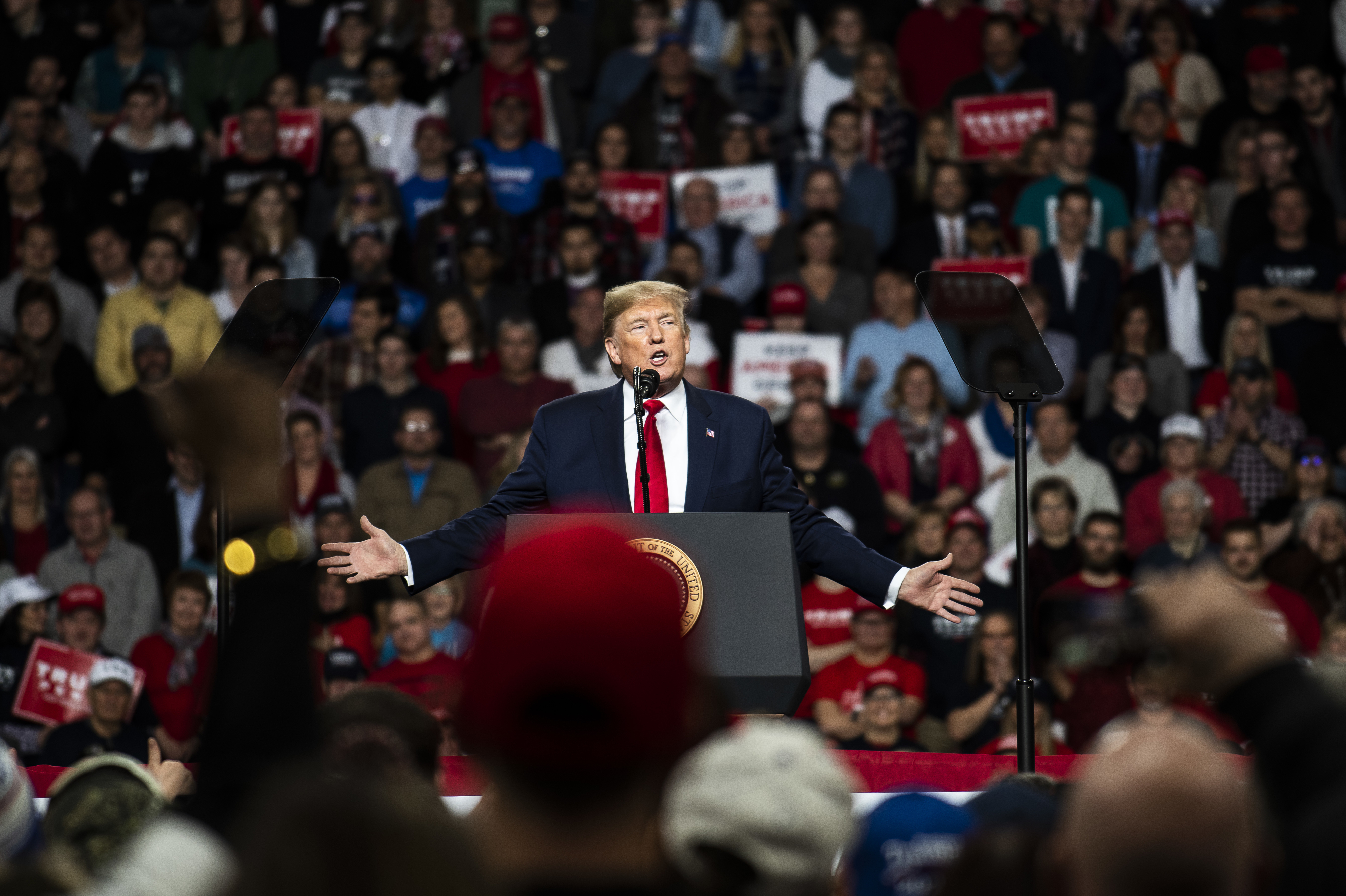 Donald Trump addresses a large crowd at a rally, standing at a podium with outstretched arms. The audience is engaged, with some holding signs