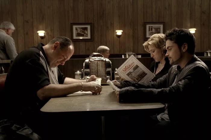 James Gandolfini, Edie Falco, and Robert Iler sit in a diner booth; James looks down, Edie reads a newspaper, and Robert looks ahead.