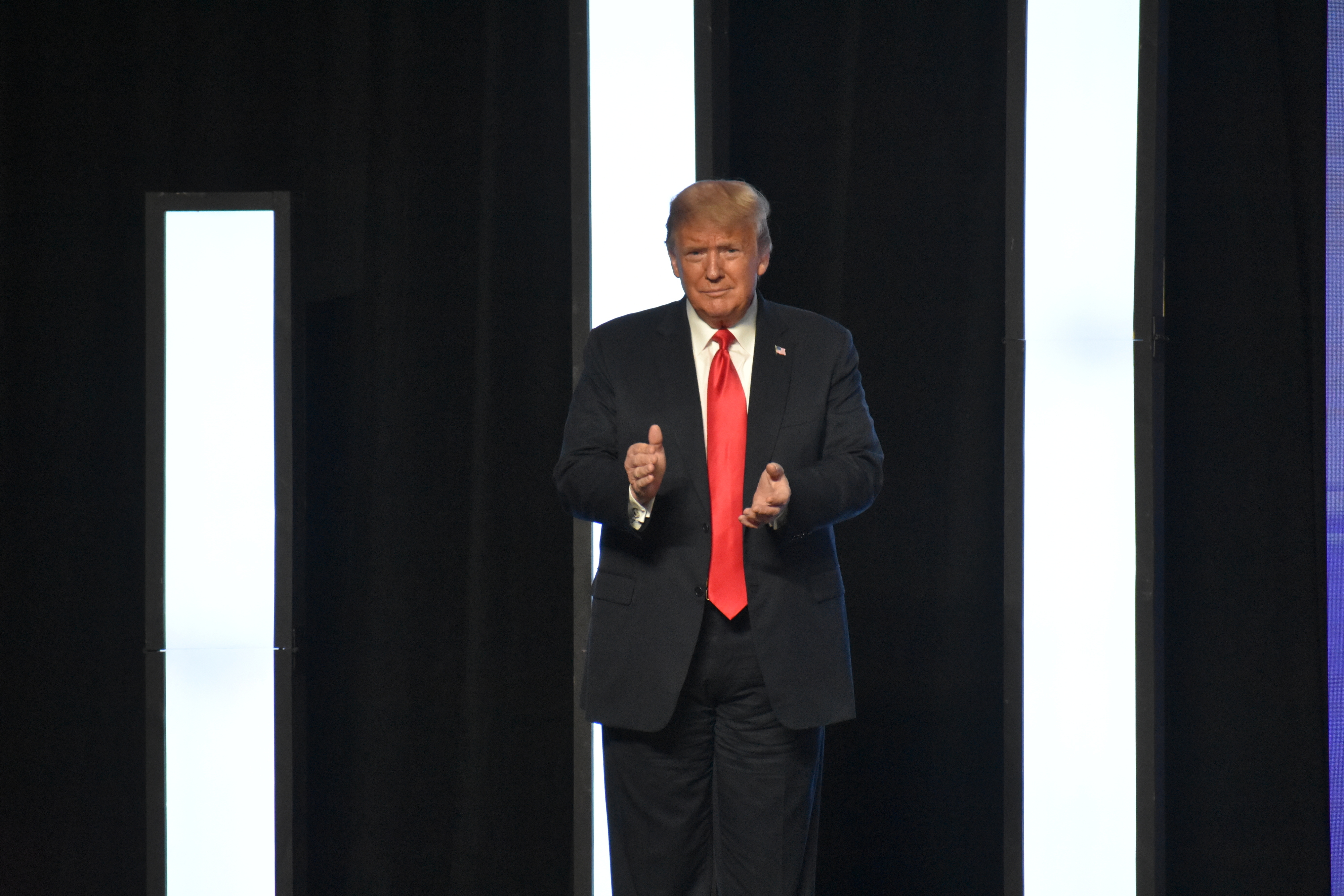 Donald Trump claps on a stage, dressed in a dark suit with a red tie, standing in front of tall, narrow light panels