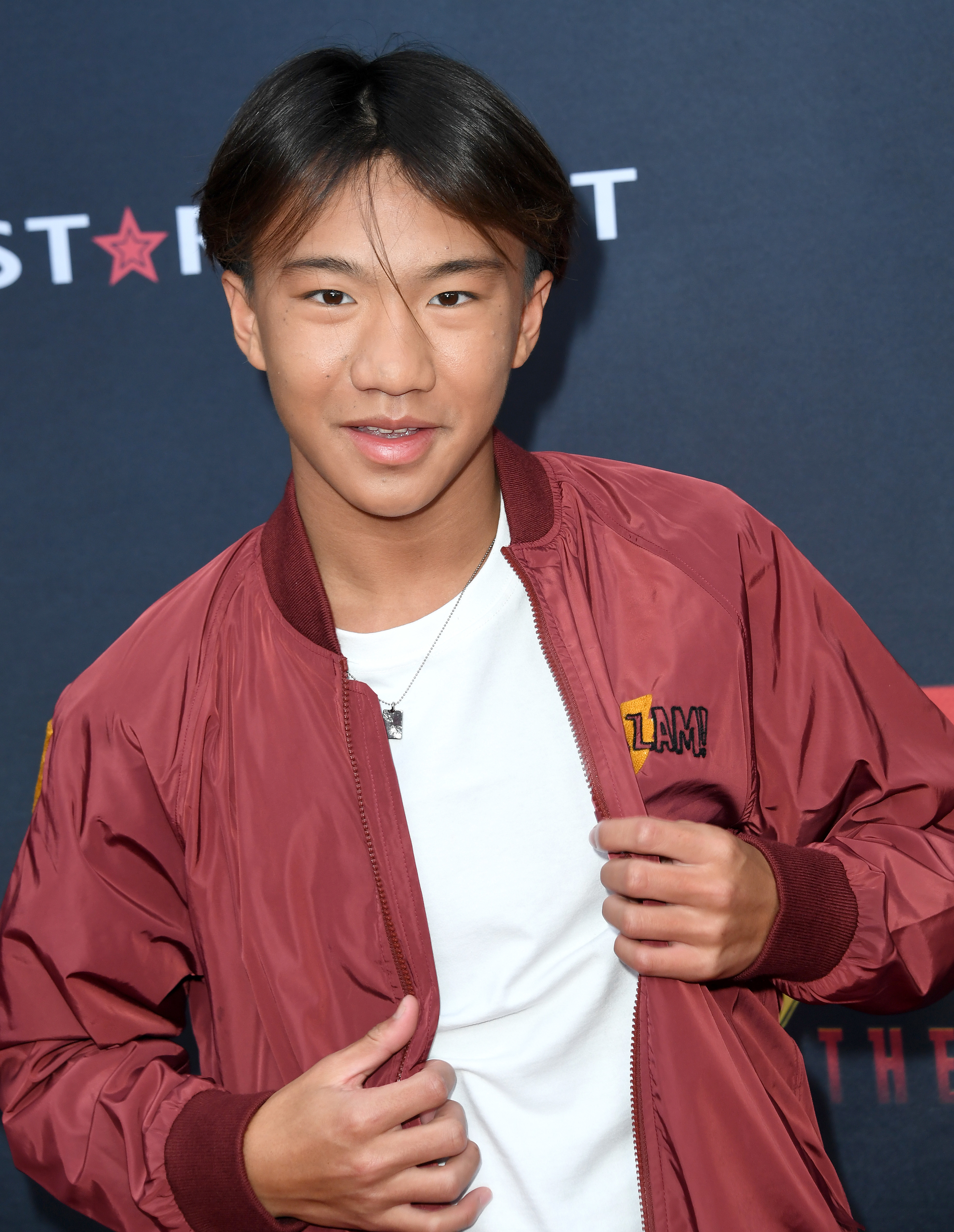 Ian Chen poses on the red carpet wearing a casual white top and maroon bomber jacket