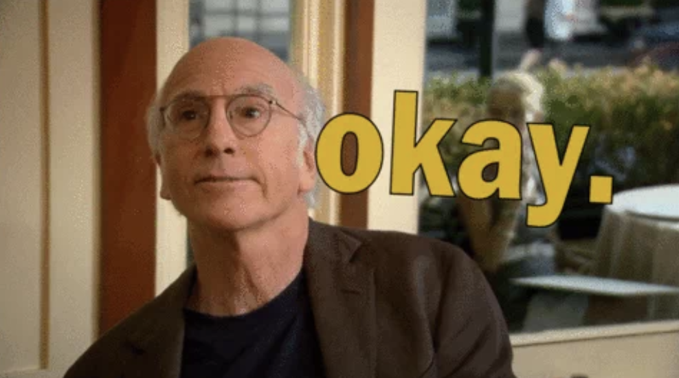 Larry David wearing glasses and a suit, with &quot;okay&quot; in large yellow text next to him
