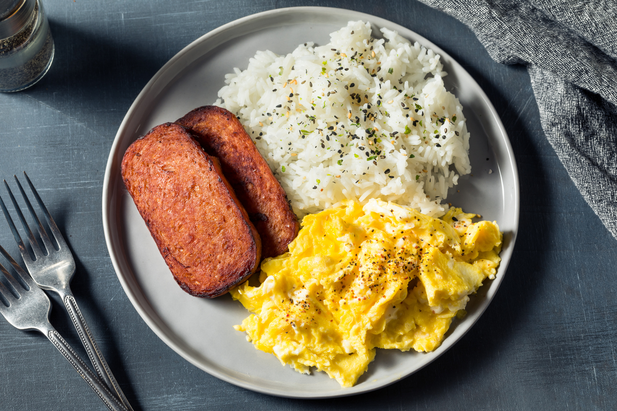 A plate of scrambled eggs, white rice sprinkled with herbs, and two slices of fried Spam, with two forks beside the plate