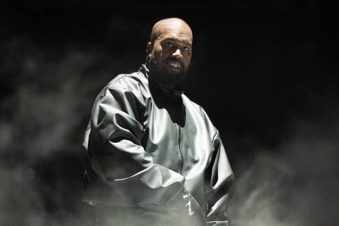 Kanye West performs on stage wearing an oversized, dark, glossy jacket. Smoke or fog surrounds the scene, creating a dramatic atmosphere
