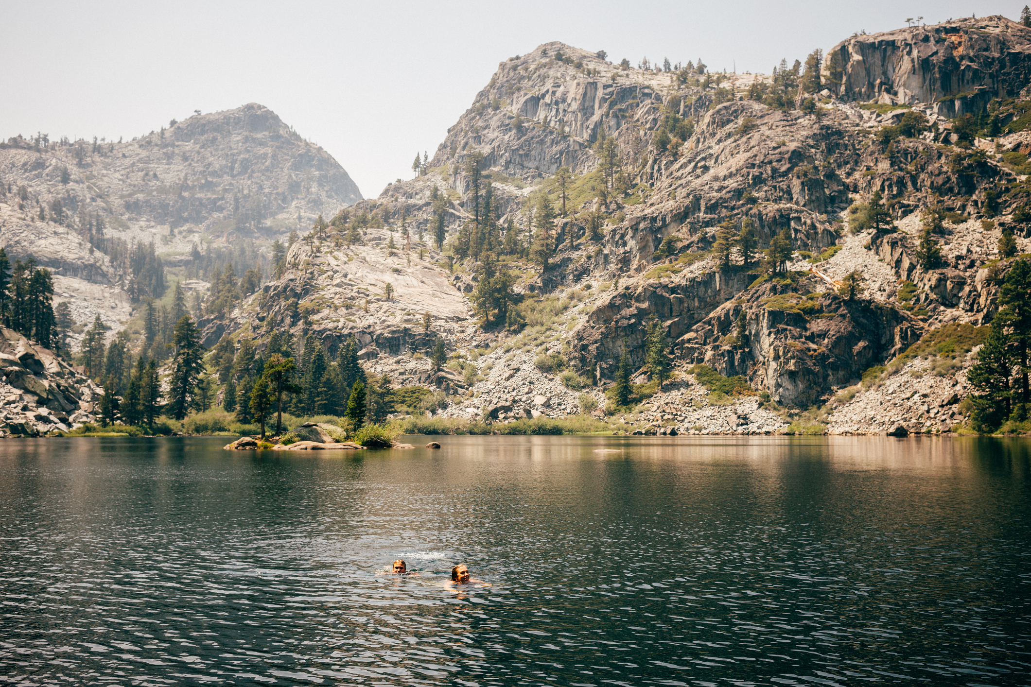 Two people swim in a serene mountain lake surrounded by rugged cliffs and forested hills