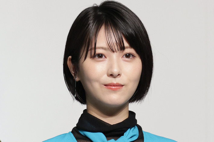 女性がカメラに向かって微笑んでいる。スタイリッシュな黒のインナーと青のアウターを着用している。