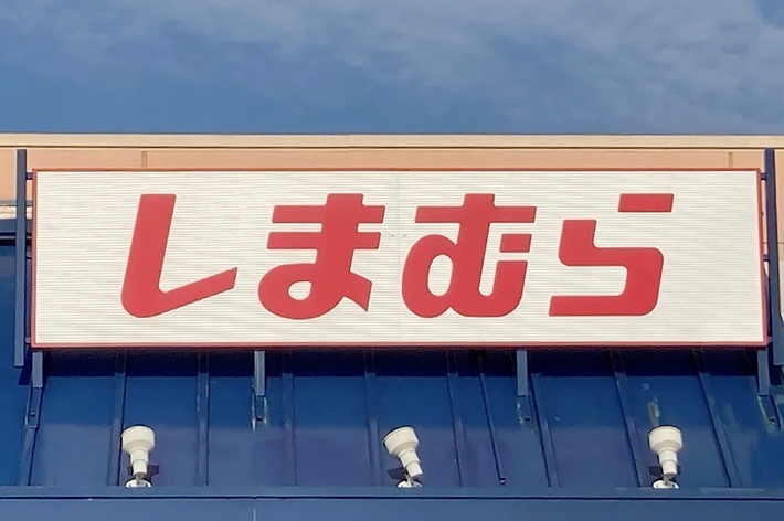 Storefront with large white sign and red Japanese Hiragana text reading &ldquo;しまむら&rdquo; (Shimamura). Several lights are below the sign