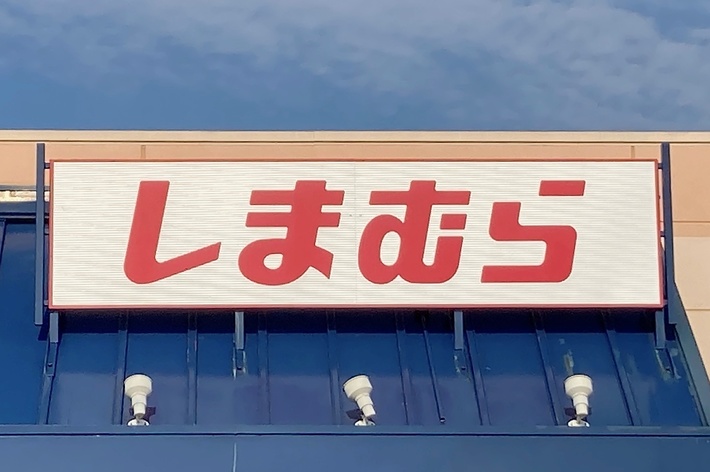 The image shows a store sign with Japanese text &ldquo;しまむら&rdquo; above the entrance. The blue sky and an accessible entrance sign are visible in the foreground
