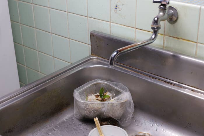流し台のシンクに古い蛇口と排水溝ネットに入った生ごみがあります。隣には箸と小さな容器が置かれています。