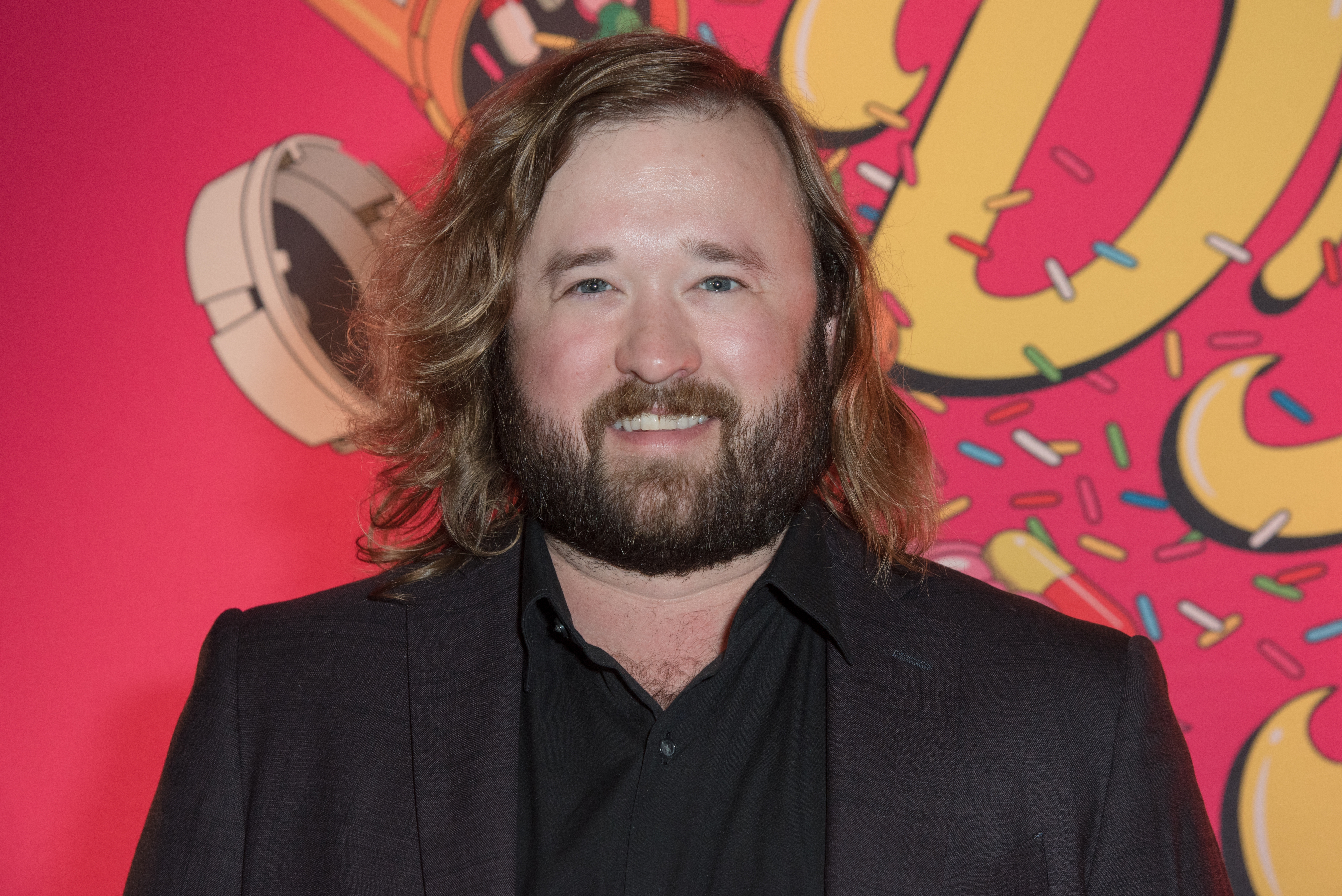 Haley Joel Osment, wearing a dark blazer over a black shirt, smiles in front of a whimsical background with colorful sprinkles