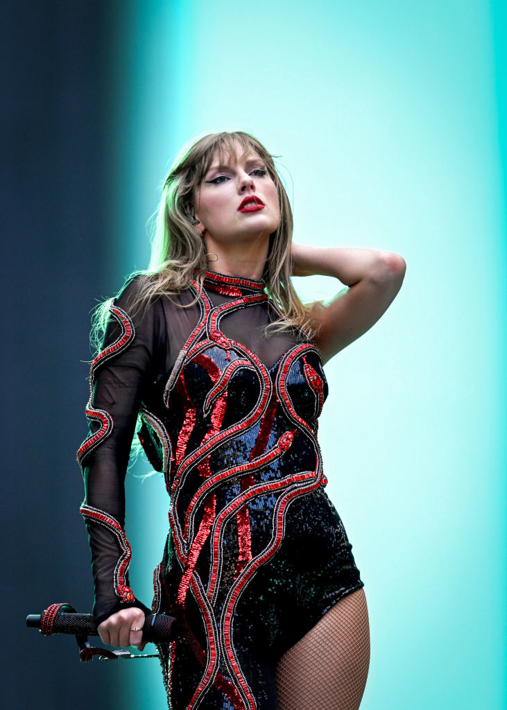 Taylor Swift performs on stage in a detailed black and red outfit, holding a microphone