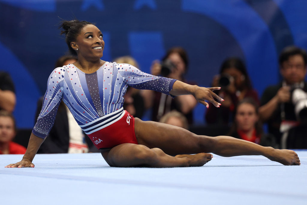 Simone Biles performs a floor routine, smiling and extending her arm while sitting on the mat at a gymnastics competition