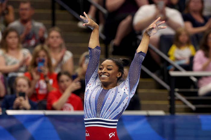 Simone Biles raises her arms successful  triumph, smiling enthusiastically, wearing a stylish gymnastics leotard with intricate patterns, successful  beforehand   of an applauding crowd