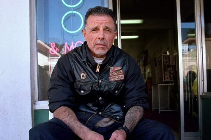 A man sits outside a shop wearing a leather jacket with patches, including one that reads "President." Neon signs are visible in the background