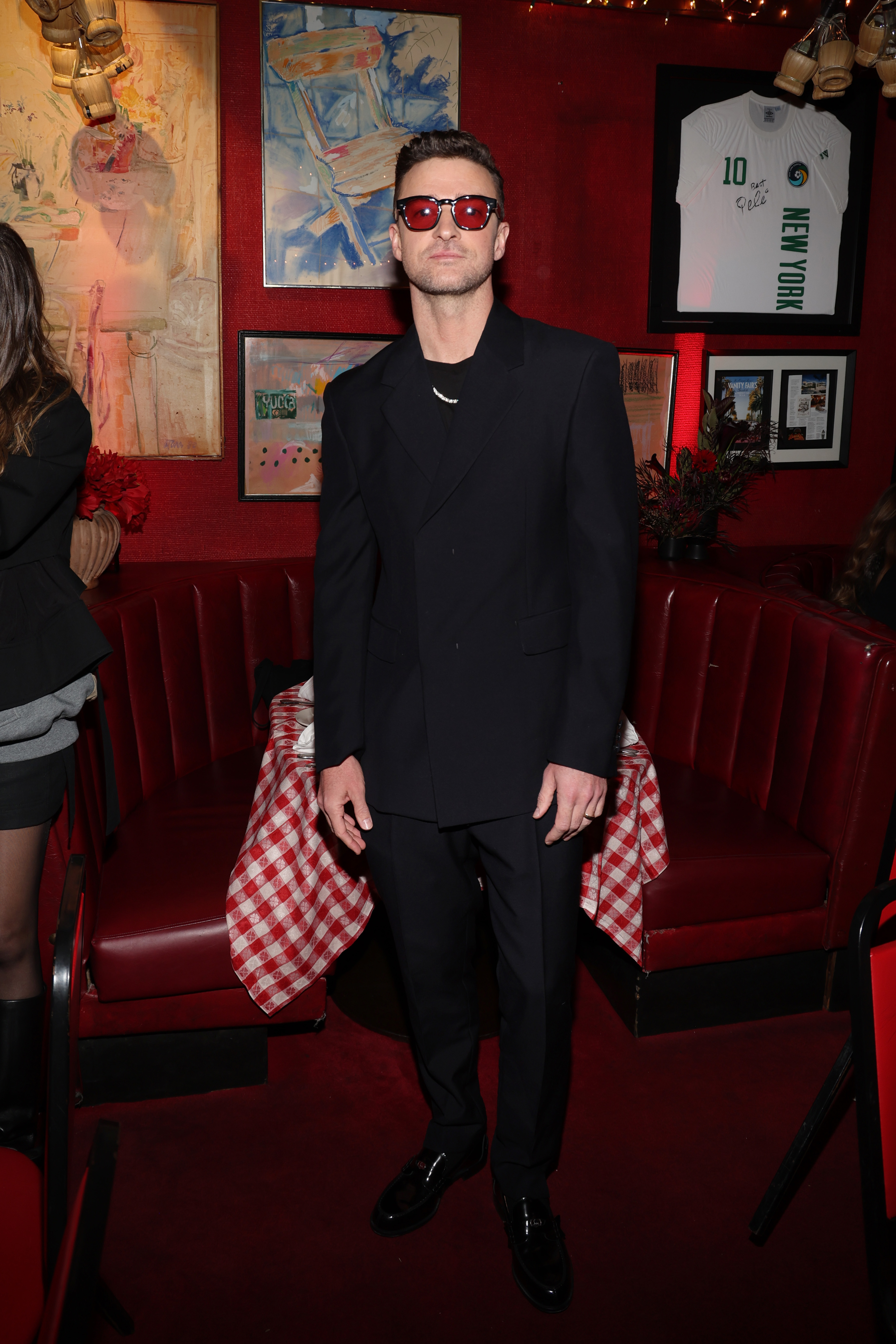 Justin Timberlake stands in a restaurant in a sleek black suit and red-tinted sunglasses. Art and memorabilia adorn the walls, with a red-checkered tablecloth visible