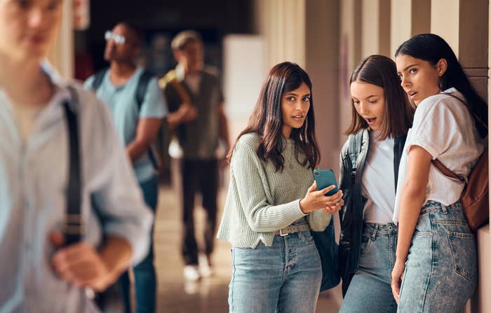 Three young women stand in a school hallway, looking at a phone together with surprised expressions. Two other students walk in the background
