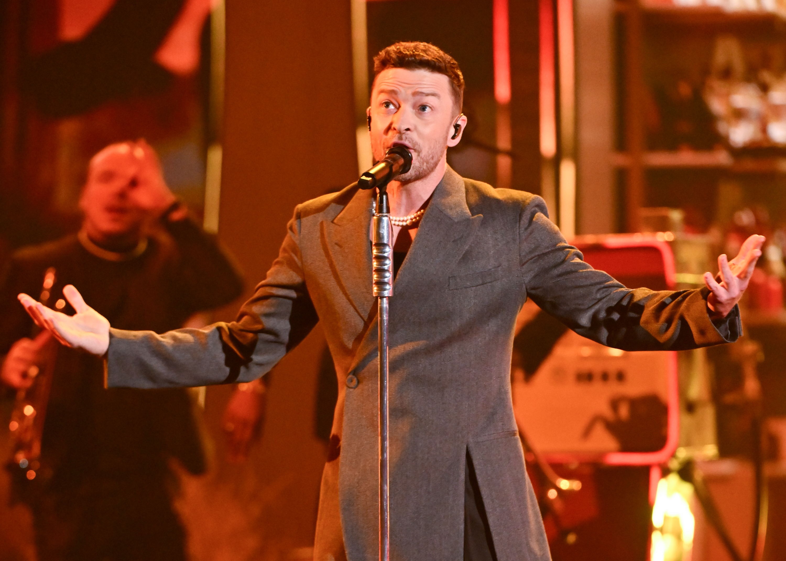 Justin Timberlake performs on stage, wearing a long, tailored coat, singing into a microphone with his arms outstretched