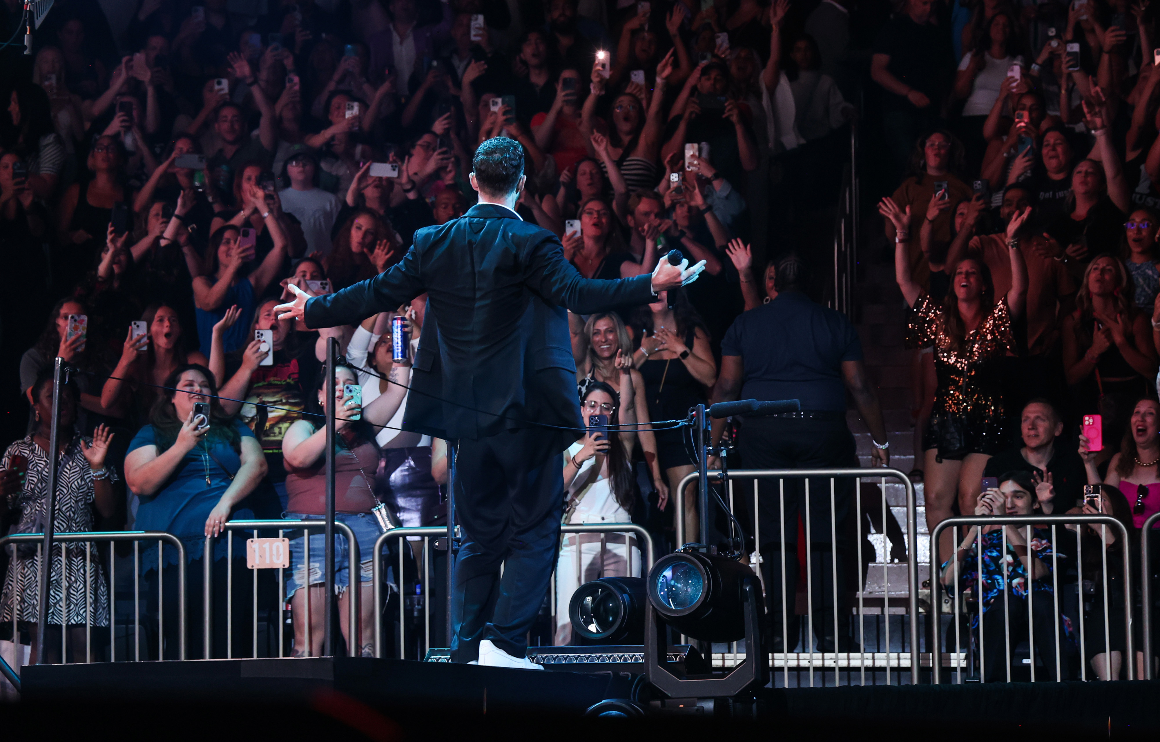 A performer with his back to the camera sings to an enthusiastic crowd, many of whom are taking photos with their phones