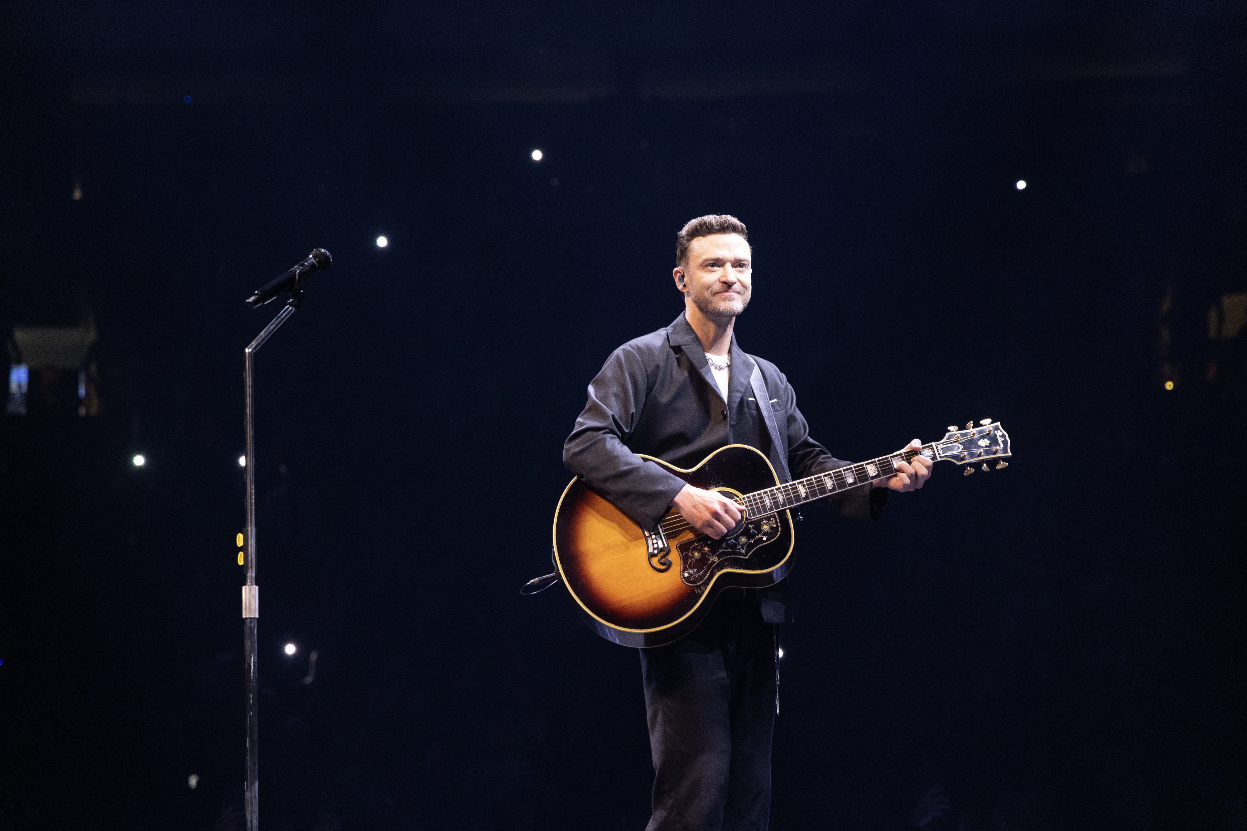 Justin Timberlake performs with an acoustic guitar on stage, wearing a dark jacket and pants, with a spotlight on him and a microphone stand nearby
