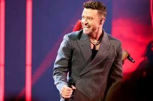 Justin Timberlake, wearing a grey blazer and black shirt, performs on stage with a microphone in hand