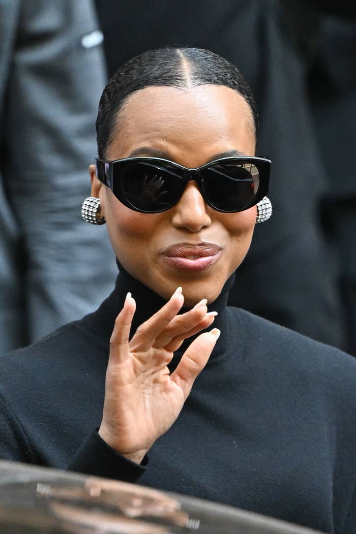Person in a black turtleneck, large black sunglasses, and checkered earrings, waving