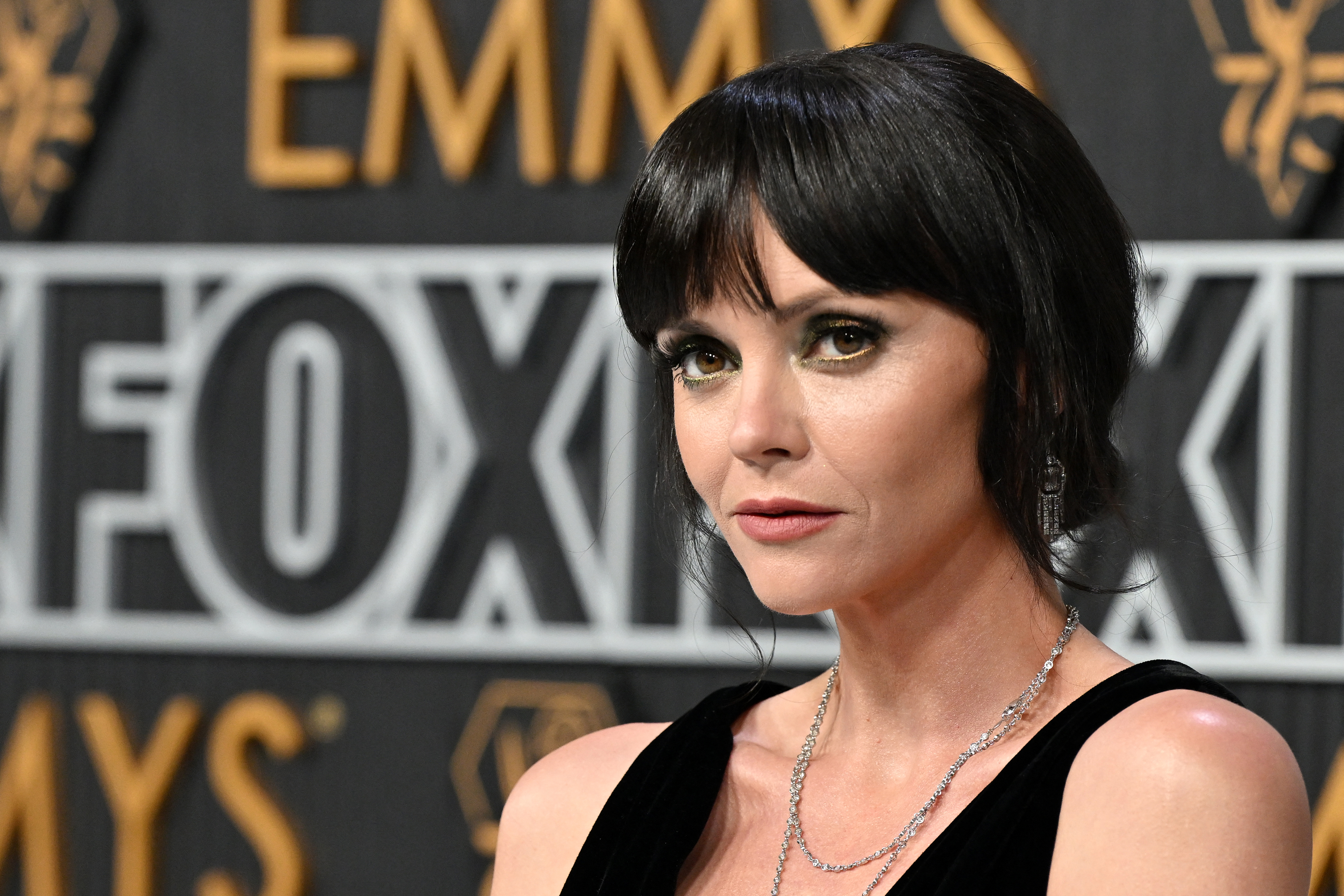 Christina Ricci poses in front of an Emmys backdrop, wearing a sleeveless black dress and silver necklace
