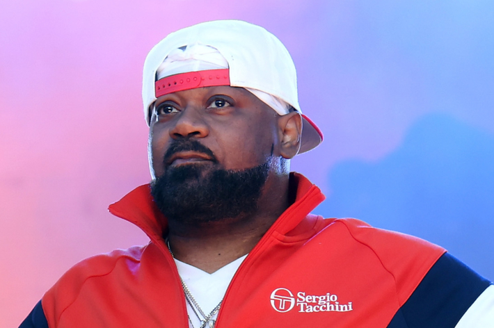 A person wearing a red and white jacket with "Sergio Tacchini" and a white and red backward cap looks into the distance against a blue and purple background