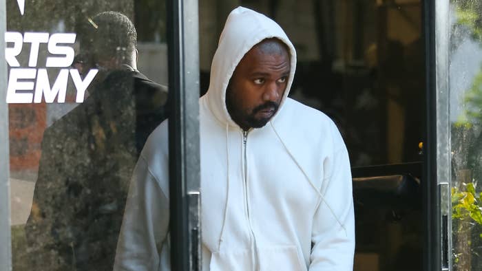 Kanye West wearing a white hoodie, standing near a glass door with a sign that reads &quot;SPORTS ACADEMY.&quot;