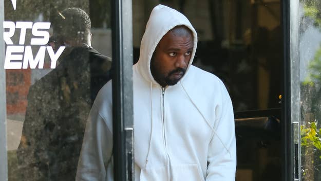 Kanye West wearing a white hoodie, standing near a glass door with a sign that reads "SPORTS ACADEMY."