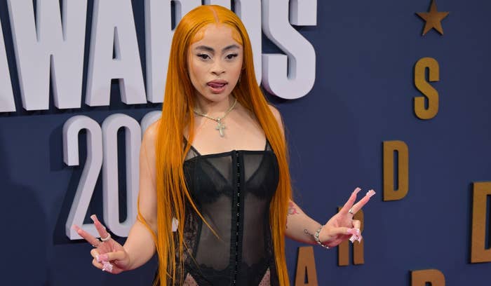 Ice Spice poses on the red carpet in a sheer black outfit, giving peace signs with both hands, at a music-related awards event where the year &quot;2023&quot; is visible