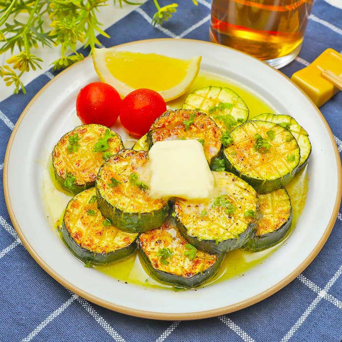 白い皿に載ったズッキーニのグリル、中央にバターが乗っている。添え物にレモンとプチトマトが付いている。