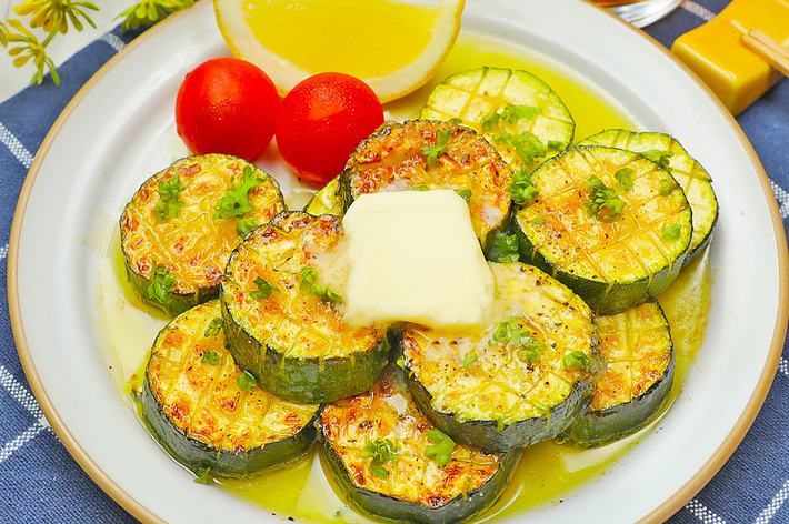 お皿に盛られた焼きズッキーニの上にバターが乗っており、トマトとレモンが添えられている食事の画像。