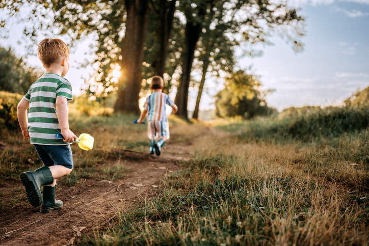 Two children playing and running on a grassy path through a forested area during sunset
