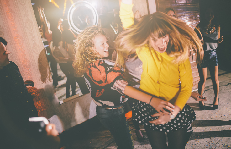 Two women dancing energetically in a lively party scene with people and lights in the background. Names unknown