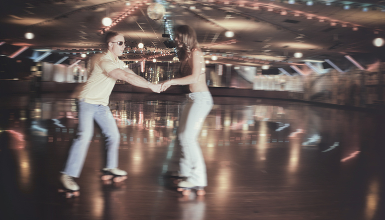 A man and woman in casual clothing roller-skate together while holding hands in a retro-style roller rink with colorful lights in the background