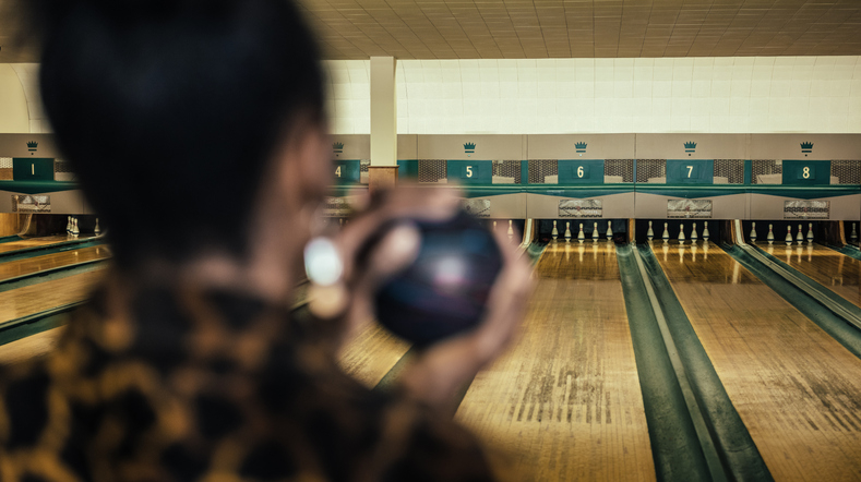 Person preparing to bowl, holding a bowling ball in a bowling alley, lanes and pins visible in the background. Names of persons unknown
