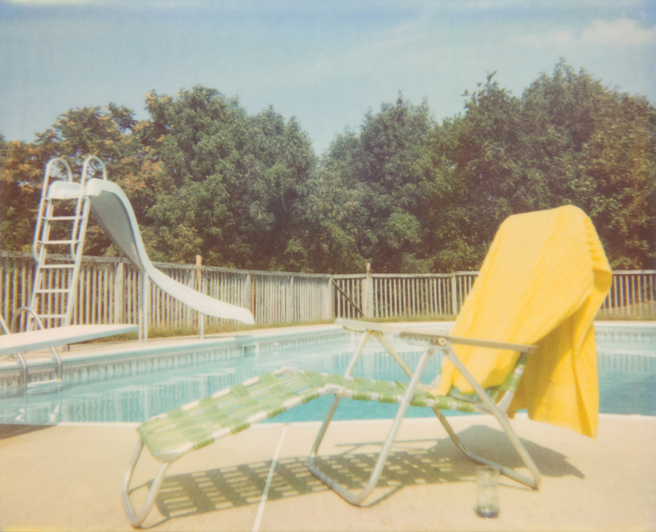 A patio chair with a yellow towel draped over it sits beside a swimming pool with a slide and diving board. Trees are visible in the background