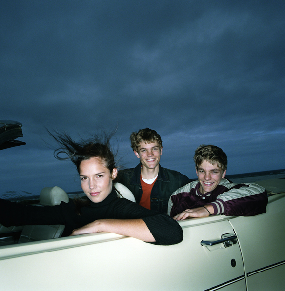 Three young people smile and pose in a convertible car with the top down. The backdrop shows a cloudy sky, creating a carefree and adventurous vibe