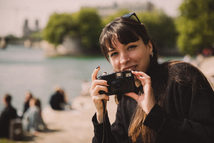 Woman with long hair and bangs holds a camera, smiling near a body of water with blurred people and greenery in the background