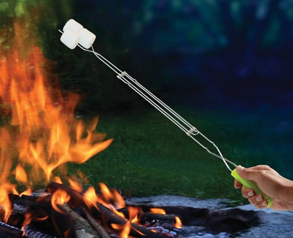 Sebuah tangan memegang tongkat pemanggang logam dengan tiga marshmallow di atas api unggun