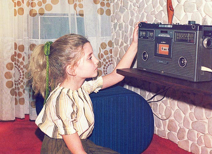 A young girl sits in front of a retro cassette player in a room with vintage decor, adjusting the device’s controls
