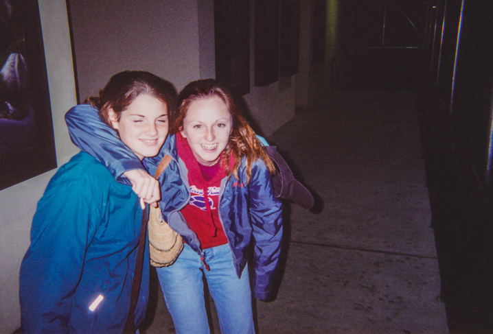 Two people smile, one with arm around the other. They are dressed casually in jackets and jeans