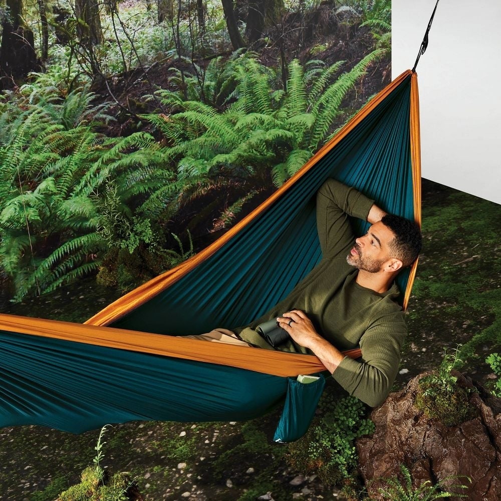 Seorang pria berbaring di tempat tidur gantung teal dan oranye di hutan